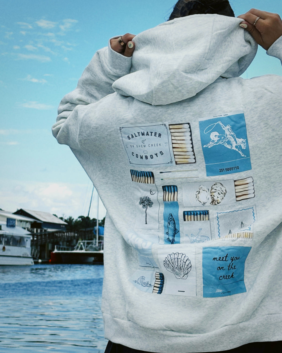 A person wearing a light gray heather hoodie with a large blue and white graphic collage print on the back, photographed outdoors by the water with boats and a dock in the background.